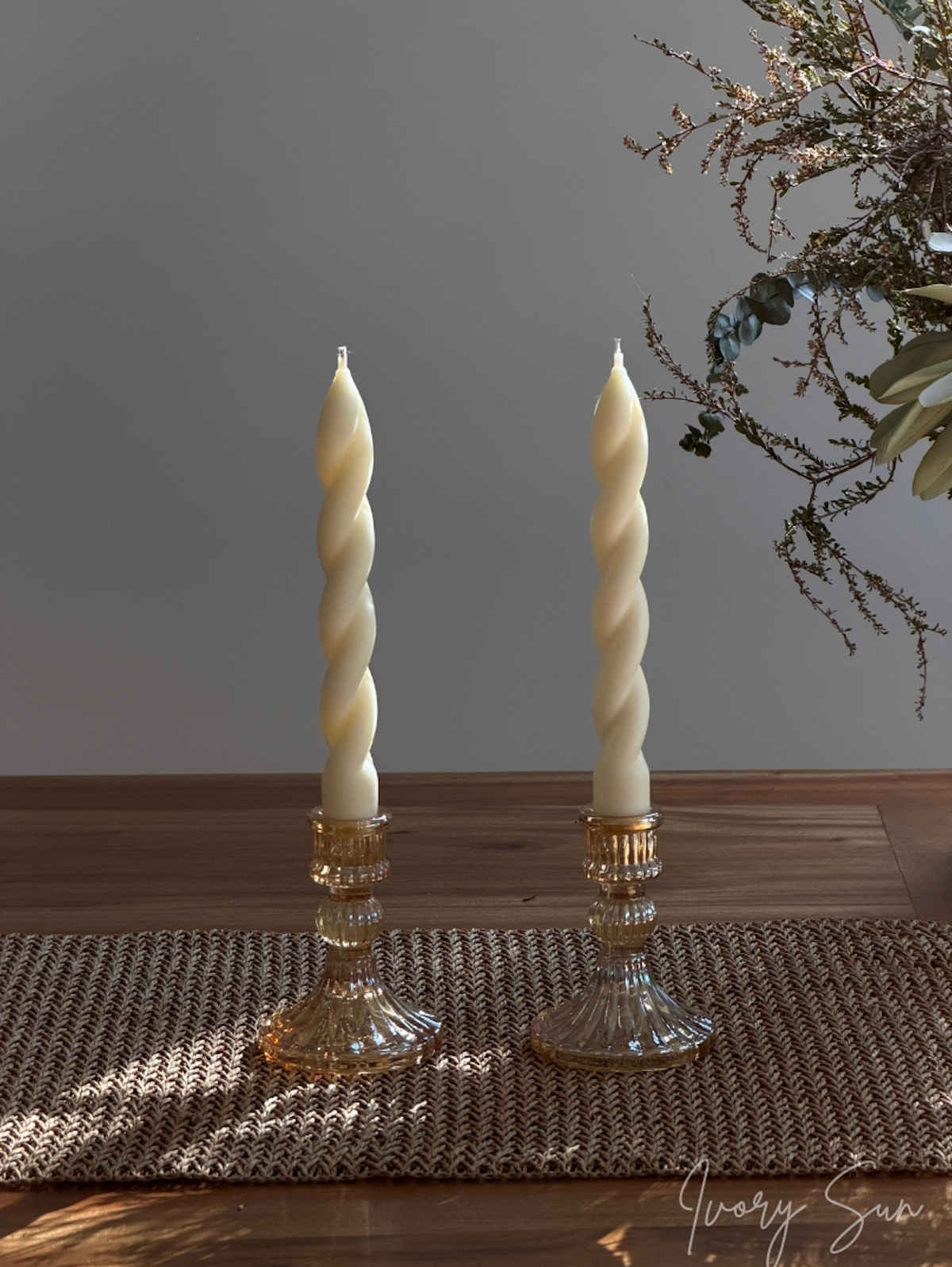 Two amber-rose coloured glass candleholders with two spiral taper candles, displayed on a woven placemat over a wooden table with a gray wall background.