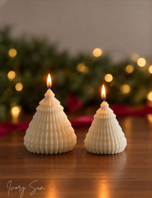 Two white ribbed Christmas tree-shaped candles with lit flames, displayed on a wooden surface with a green garland and warm string lights in the background. The brand name 'Ivory Sun' is visible in the bottom left corner. Sculptural Christmas Tree Candles handmade by Ivory Sun.