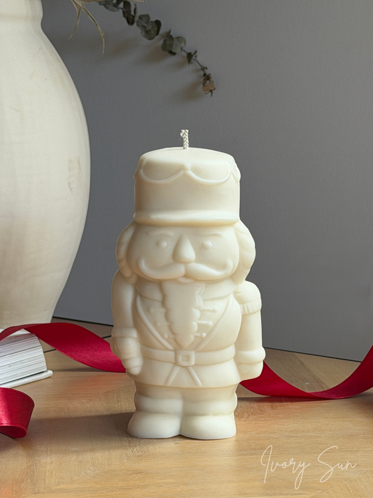 A cream soy Nutcracker candle sits on a wooden surface, styled with a red ribbon, a large white ceramic vase, and a gray background wall. Handmade Christmas candles by Ivory Sun, beautifully presented as a Christmas gift set. Discover Christmas gift ideas and Christmas Candles in Australia.