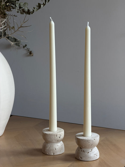 Two tall cream taper candles standing in round, textured stone candle holders on a wooden surface against a gray wall, with a white vase and dried branches partially visible at the left edge. Handmade taper candles by Ivory Sun.