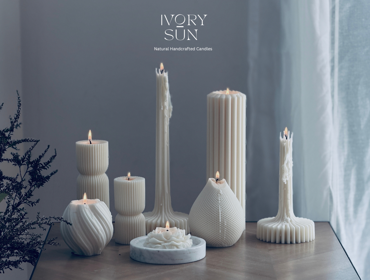 Candle gift cards. A collection of white handcrafted soy candles in various sculptural shapes and heights, styled on a wooden table against a gray wall with a curtain. The brand name 'IVORY SUN' is displayed at the top. Ivory Sun gift cards available online.