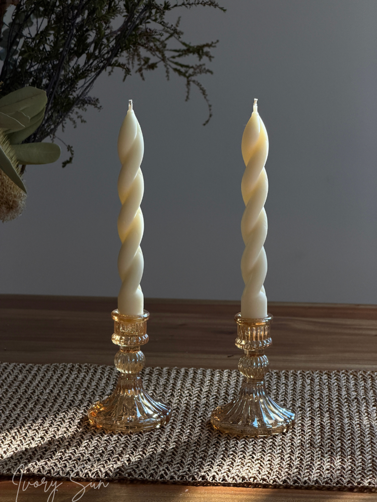 Two amber-rose coloured glass candle holders with white soy wax twisted taper candles, placed on a woven placemat on a wooden table with a gray wall background. Handmade candles by Ivory Sun.