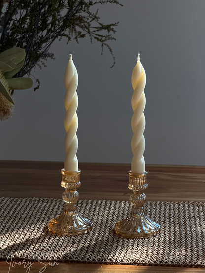 Two amber-rose coloured glass candle holders with white soy wax twisted taper candles, placed on a woven placemat on a wooden table with a gray wall background. Handmade candles by Ivory Sun.