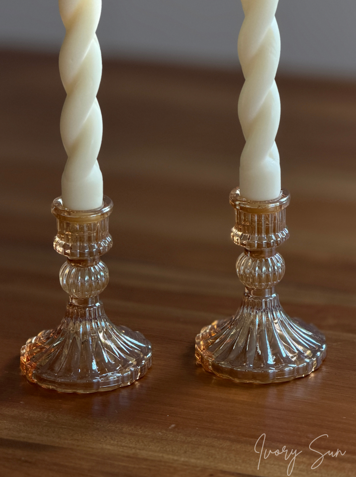 Two amber-rose glass candle holders paired with white soy wax twisted taper candles, on a wooden table against a soft grey wall. Elegant handmade candles by Ivory Sun, perfect for creating a warm and inviting atmosphere.