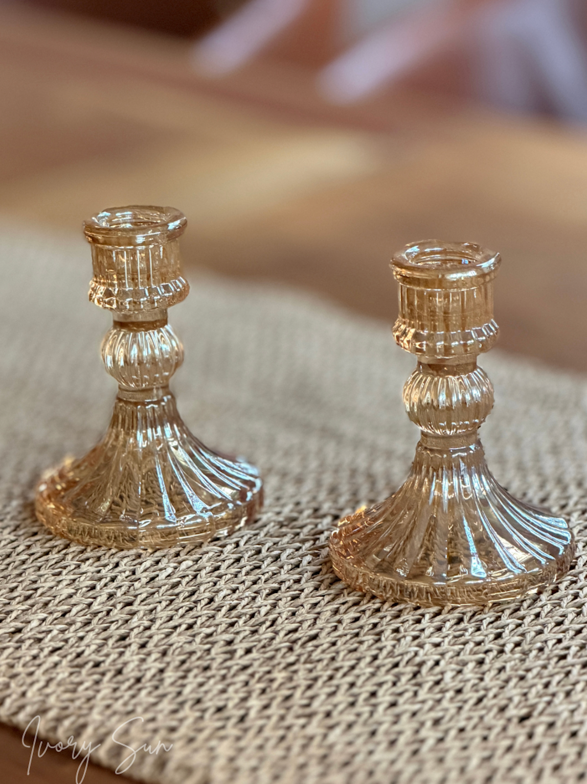 Two small ribbed glass candle holders in a warm amber-tinted finish, displayed on a woven mat surface with a softly blurred wooden background.