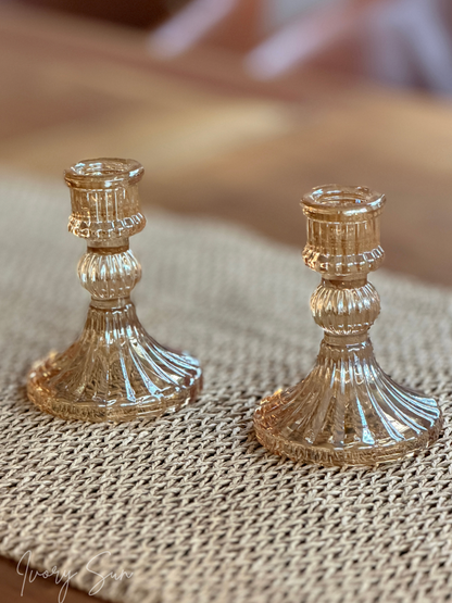 Two small ribbed glass candle holders in a warm amber-tinted finish, displayed on a woven mat surface with a softly blurred wooden background.