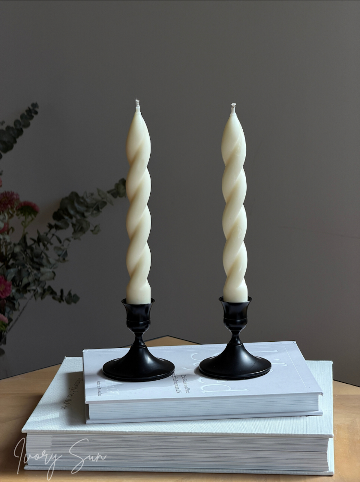 Two twisted taper candles displayed in elegant black candle holders Australia on stacked books with a soft floral background. These metal candle holders feature a modern design, perfect for styling taper candles, dining tables and special occasions. The Ivory Sun candle holder collection includes stylish candle accessories, twin candle holders, wedding candle holders, dinner party candle holders, decorative candle holders set and timeless table décor pieces for table styling. 