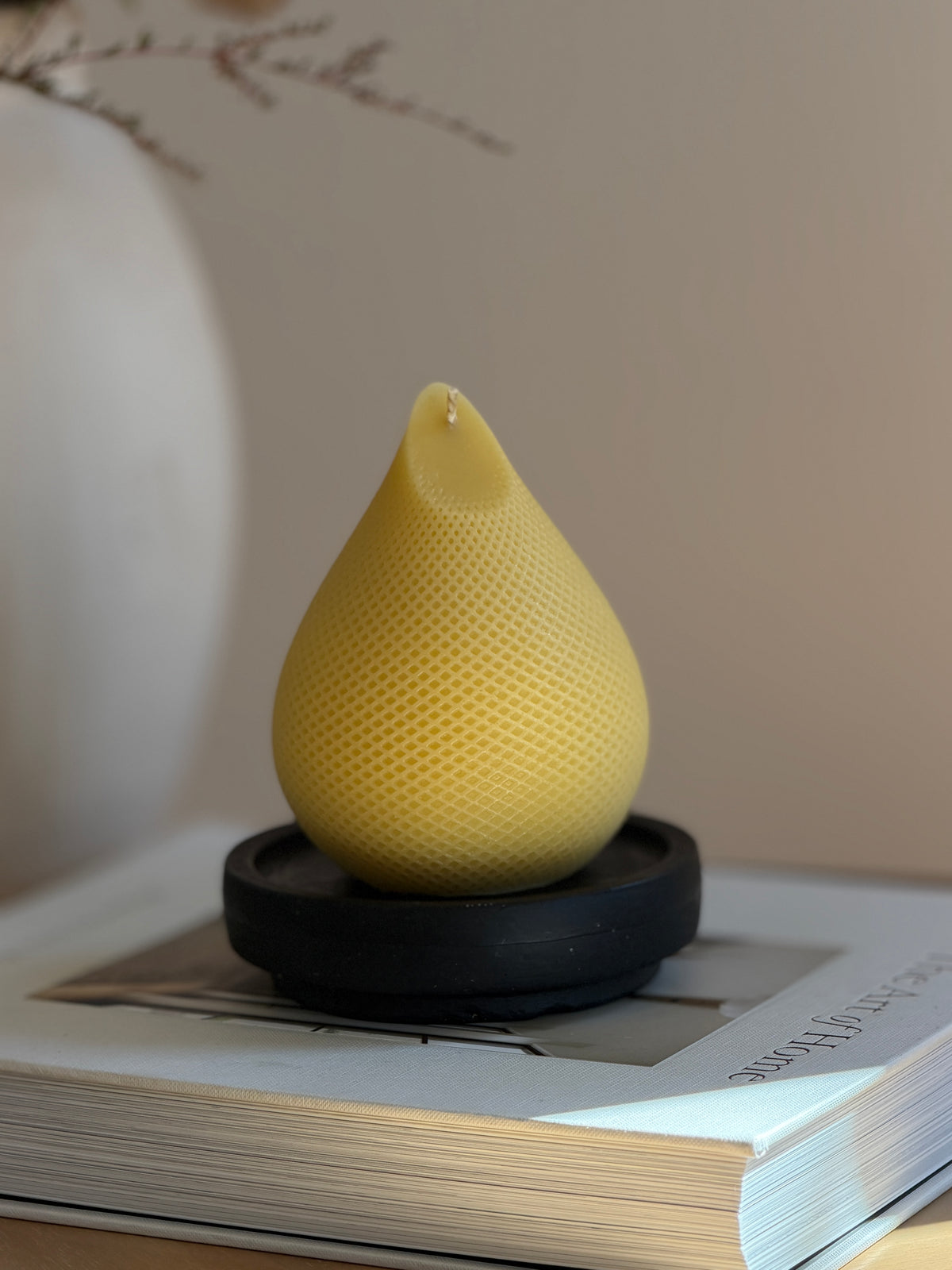 A caramel beeswax teardrop-shaped candle with a textured honeycomb pattern sits on a black candle tray, placed on top of a book with a soft-focus neutral background. Handmade by Ivory Sun.