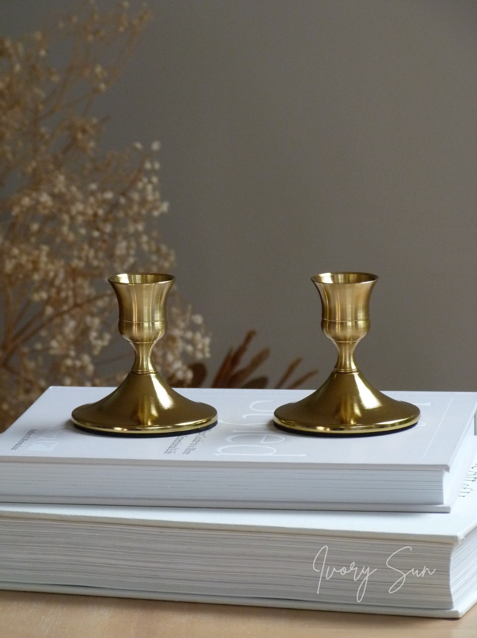 Two gold candle holders for taper candles on a stack of books with a neutral background.
metal candle holders, gold candle holders, golden candle holders, candle holders australia, candle holder, candle holder australia, ivory sun candle holder, candle accessories, wedding candle holders, dinner party candle holders, decor candle holders, twin candle holders, table decor