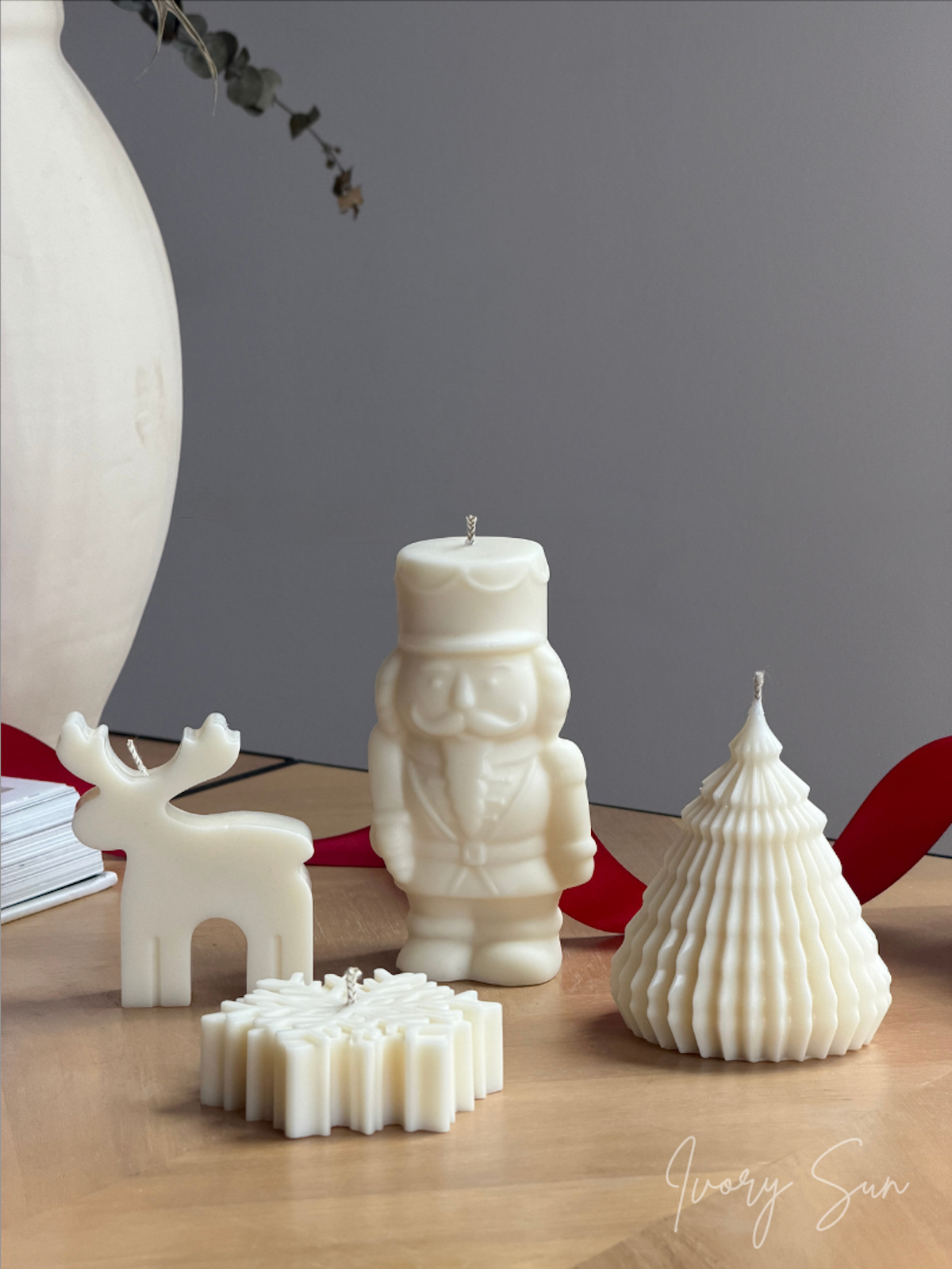 A collection of white sculptural candles displayed on a light wood table against a gray background, including a nutcracker, a reindeer, a Christmas tree, and a snowflake design, with a red ribbon and a large white vase in the background. Handmade Christmas candles by Ivory Sun, beautifully presented as a Christmas gift set. Discover Christmas gift ideas and Christmas Candles in Australia.
