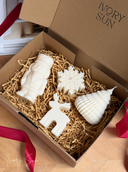 An open gift box filled with shredded paper and four white Christmas themed candles shaped like Nutcracker, a snowflake, a reindeer and a Christmas tree. The brand name 'IVORY SUN' is printed on the inside of the box lid, and a red ribbon is tied around the box. Handmade Christmas candles by Ivory Sun, beautifully presented as a Christmas gift set. Discover Christmas gift ideas and Christmas Candles in Australia.