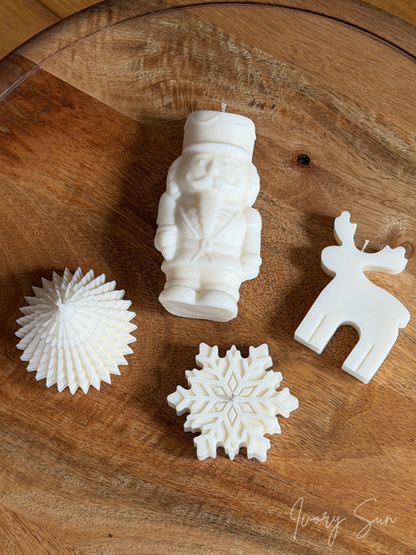 Four white Christmas themed candles arranged on a round wooden table: a nutcracker candle, a reindeer candle, a snowflake candle, and a Christmas Tree candle. The brand name 'Ivory Sun' appears in the bottom right corner. Handmade Christmas candles by Ivory Sun, beautifully presented as a Christmas gift set. Discover Christmas gift ideas and Christmas Candles in Australia.