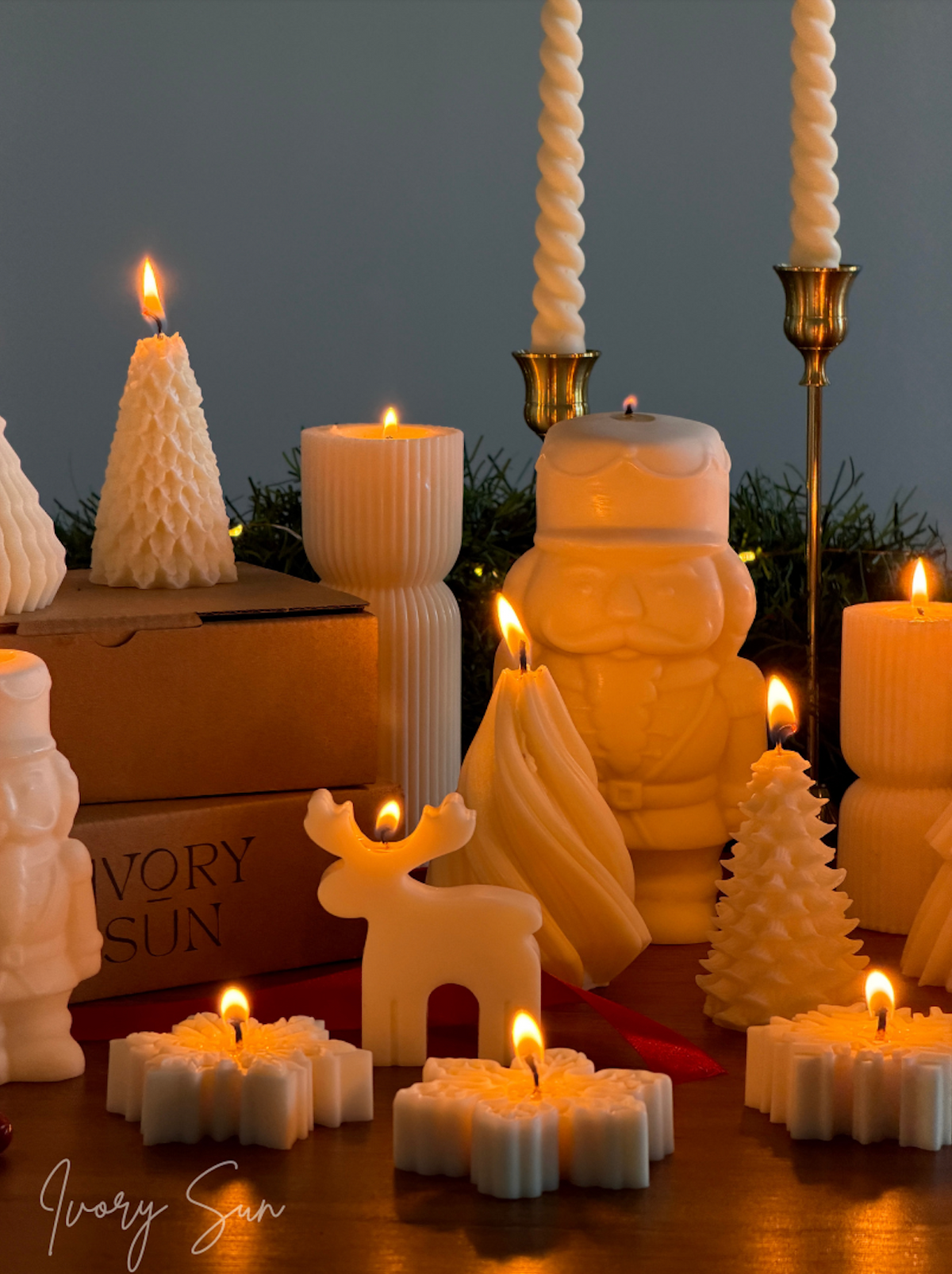 A collection of white Chritmas candles from Ivory Sun, including Santa Claus, reindeer, Christmas trees, and twisted pillar candles, arranged on a wooden surface with lit flames against a dark background. Handmade by Ivory Sun.