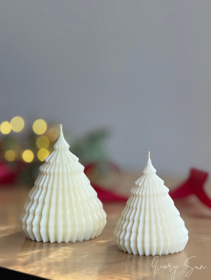 Two white soy ribbed Christmas tree-shaped candles on a wooden surface with a soft gray background and blurred holiday lights. Sculptural Christmas Tree Candles handmade by Ivory Sun.