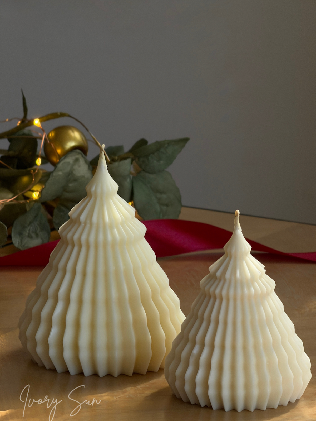 Two ribbed, tree-shaped candles in an ivory/cream colour, styled on a wooden surface with green leaves, gold ornaments, and a red ribbon in the background. Sculptural Christmas Tree Candles handmade by Ivory Sun.