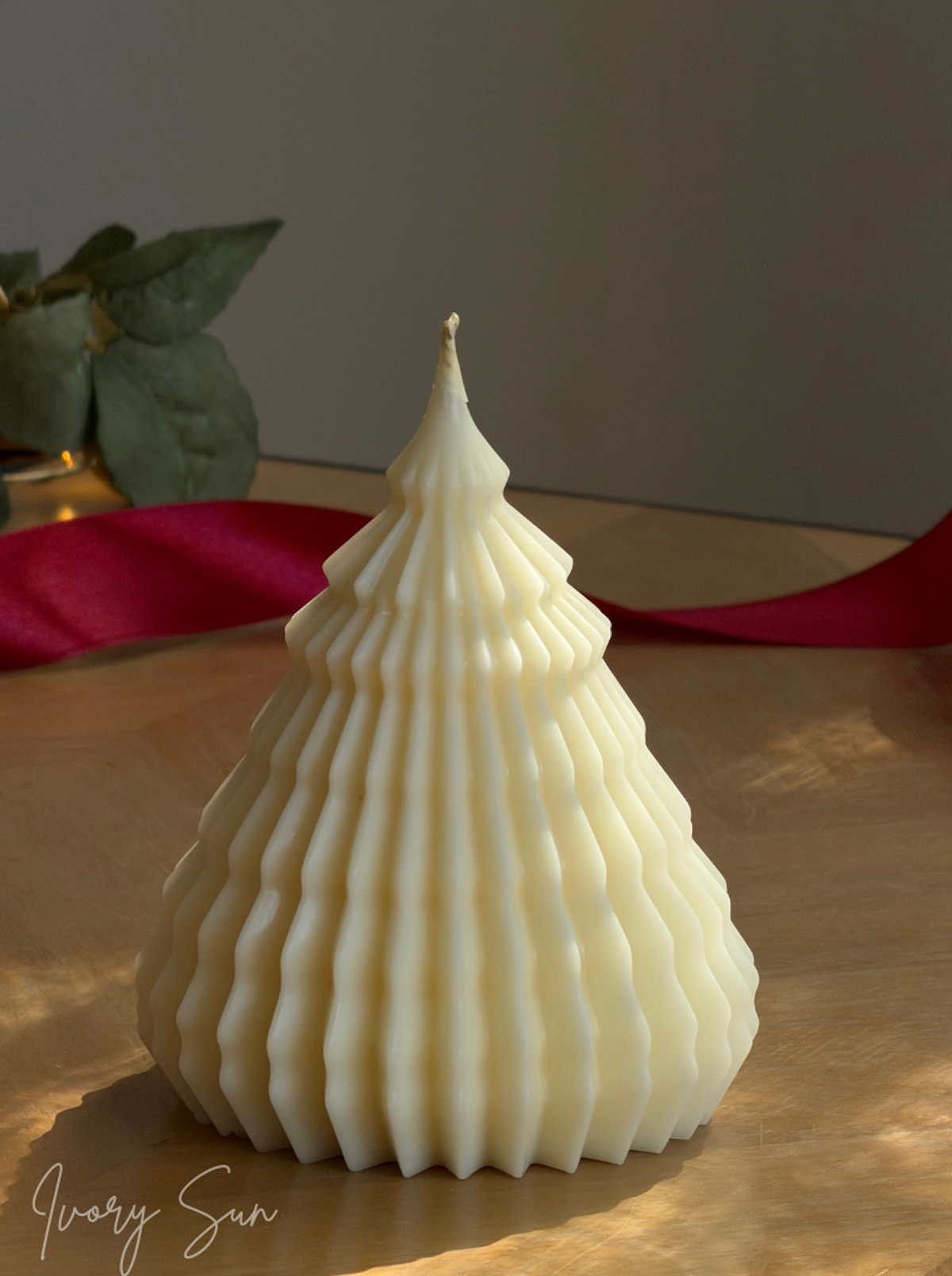 An ivory tree-shaped candle with vertical fluted ridges, displayed on a wooden surface with a red ribbon in the background. Sculptural Christmas Tree Candles handmade by Ivory Sun. 