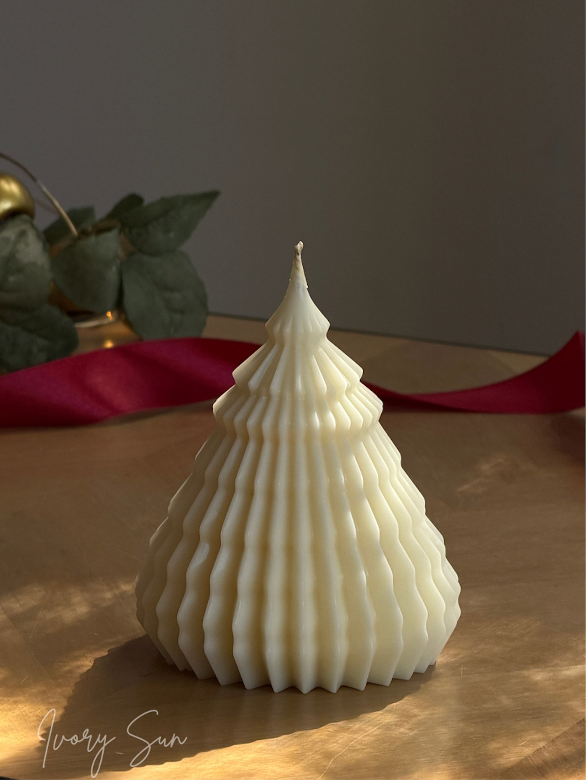 An off-white, ribbed Christmas tree-shaped candle sits on a wooden surface with a red ribbon in the background. Sculptural Christmas Tree Candles handmade by Ivory Sun.