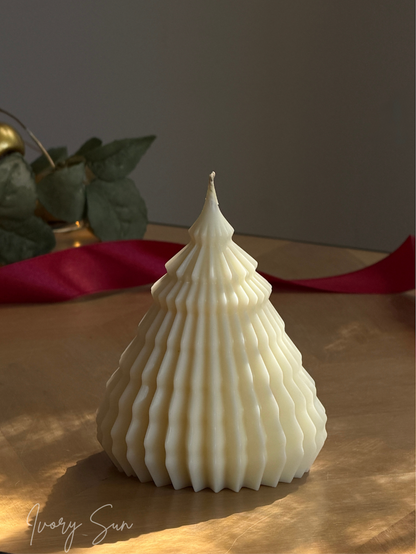 An off-white, ribbed Christmas tree-shaped candle sits on a wooden surface with a red ribbon in the background. Sculptural Christmas Tree Candles handmade by Ivory Sun.