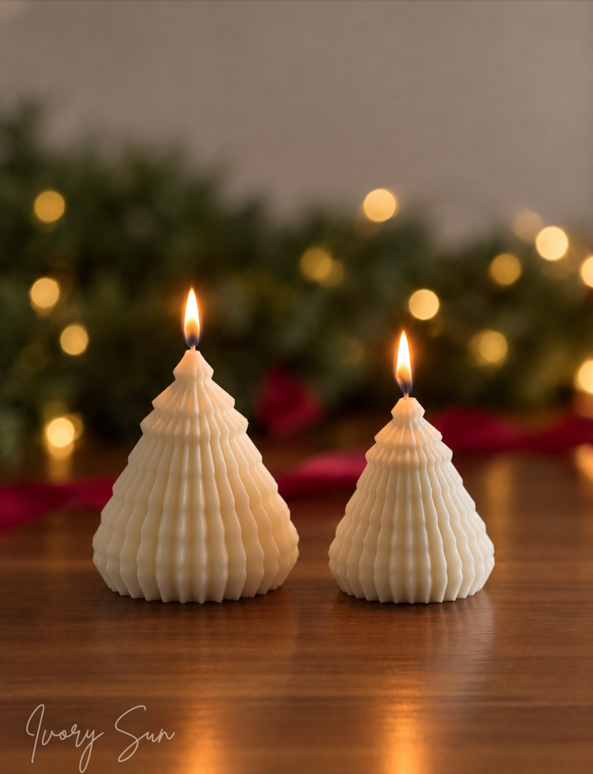 Two white ribbed Christmas tree-shaped candles with lit flames, displayed on a wooden surface with a green garland and warm string lights in the background. The brand name 'Ivory Sun' is visible in the bottom left corner. Sculptural Christmas Tree Candles handmade by Ivory Sun.