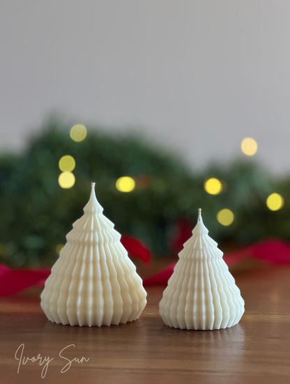Two small ribbed, tree-shaped white soy candles standing on a wooden surface, with a blurred holiday greenery and warm string lights in the background. The brand name 'Ivory Sun' is written in white script in the bottom-left corner. Sculptural Christmas Tree Candles handmade by Ivory Sun.