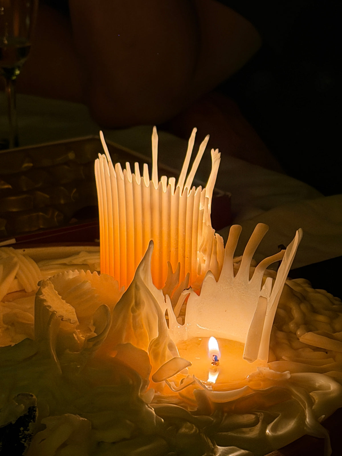 A decorative soy wax candle in a sculpted, organic shape. Melted pillar candles forming a tall cluster of white wax wicks rising from the back, all illuminated by warm candlelight.