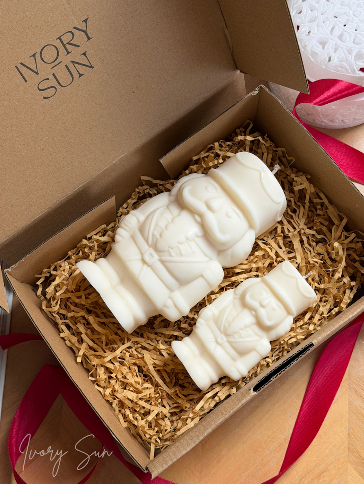 Two ivory white Nutcracker candles displayed inside a brown cardboard gift box filled with shredded paper, with the brand name 'Ivory Sun' printed on the inside lid and a red ribbon nearby. Handmade Christmas candles by Ivory Sun, beautifully presented as a Christmas gift set. Discover Christmas gift ideas and Christmas Candles in Australia.