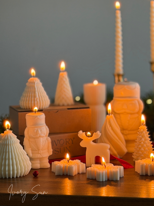 A collection of white Christmas soy wax candles shaped like a nutcracker, reindeer, Christmas trees, snowflakes,  arranged on a wooden table with warm candlelight against a soft gray background. Handmade Christmas candles by Ivory Sun.