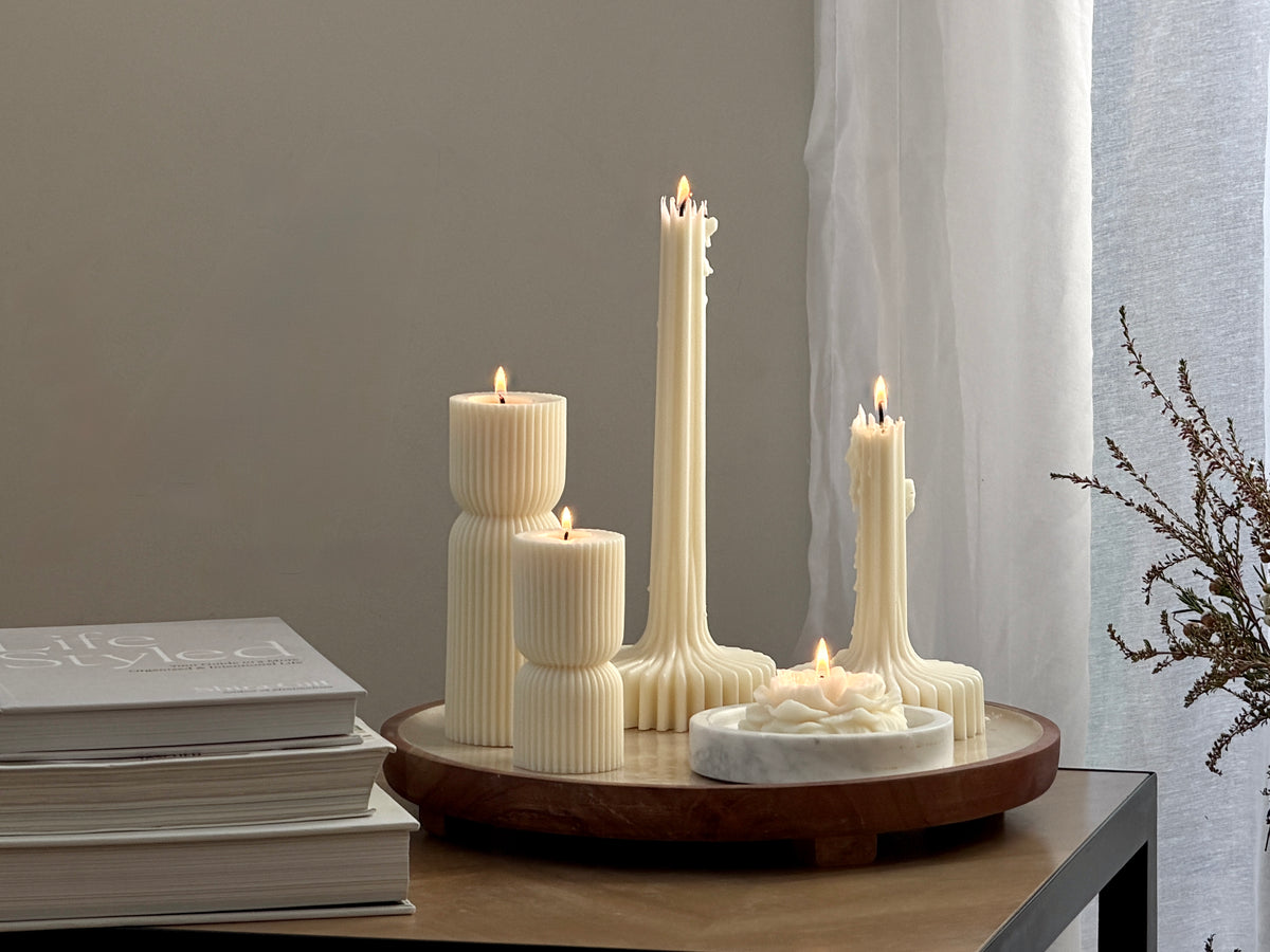 A set of cream-colored ribbed pillar and taper candles arranged on a round wooden tray, with one small candle in a white marble holder, styled on a wooden table next to books and sheer curtains. Handmade by Ivory Sun.