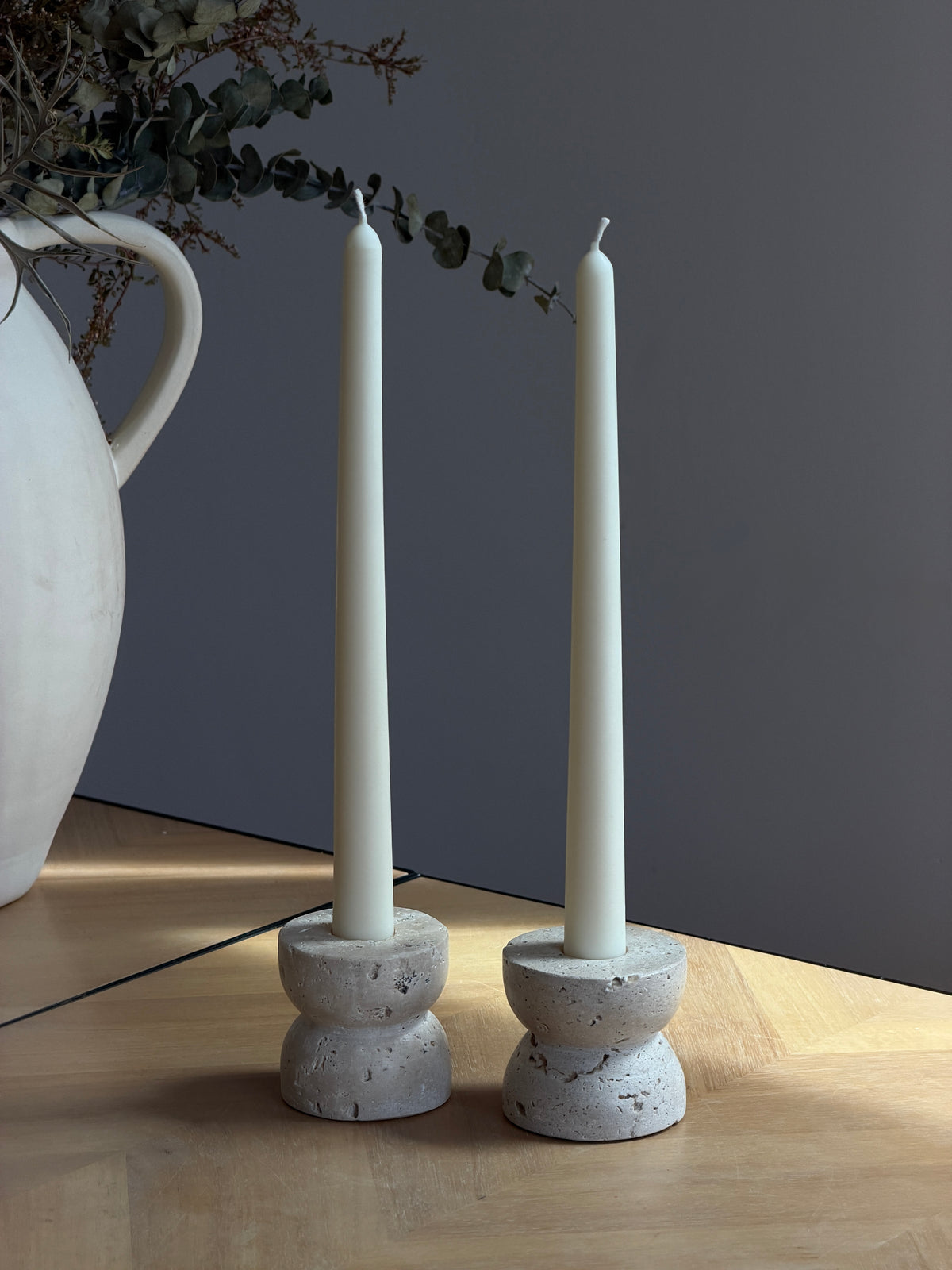 Two tall white taper candles in round textured stone holders, placed on a light wood tabletop against a dark gray wall, with a white ceramic pitcher and eucalyptus visible at the left edge. Handmade taper candles by Ivory Sun.