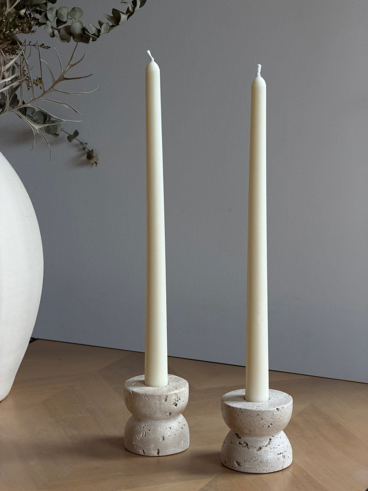 Two tall cream taper candles standing in round, textured stone candle holders on a wooden surface against a gray wall, with a white vase and dried branches partially visible at the left edge. Handmade taper candles by Ivory Sun.