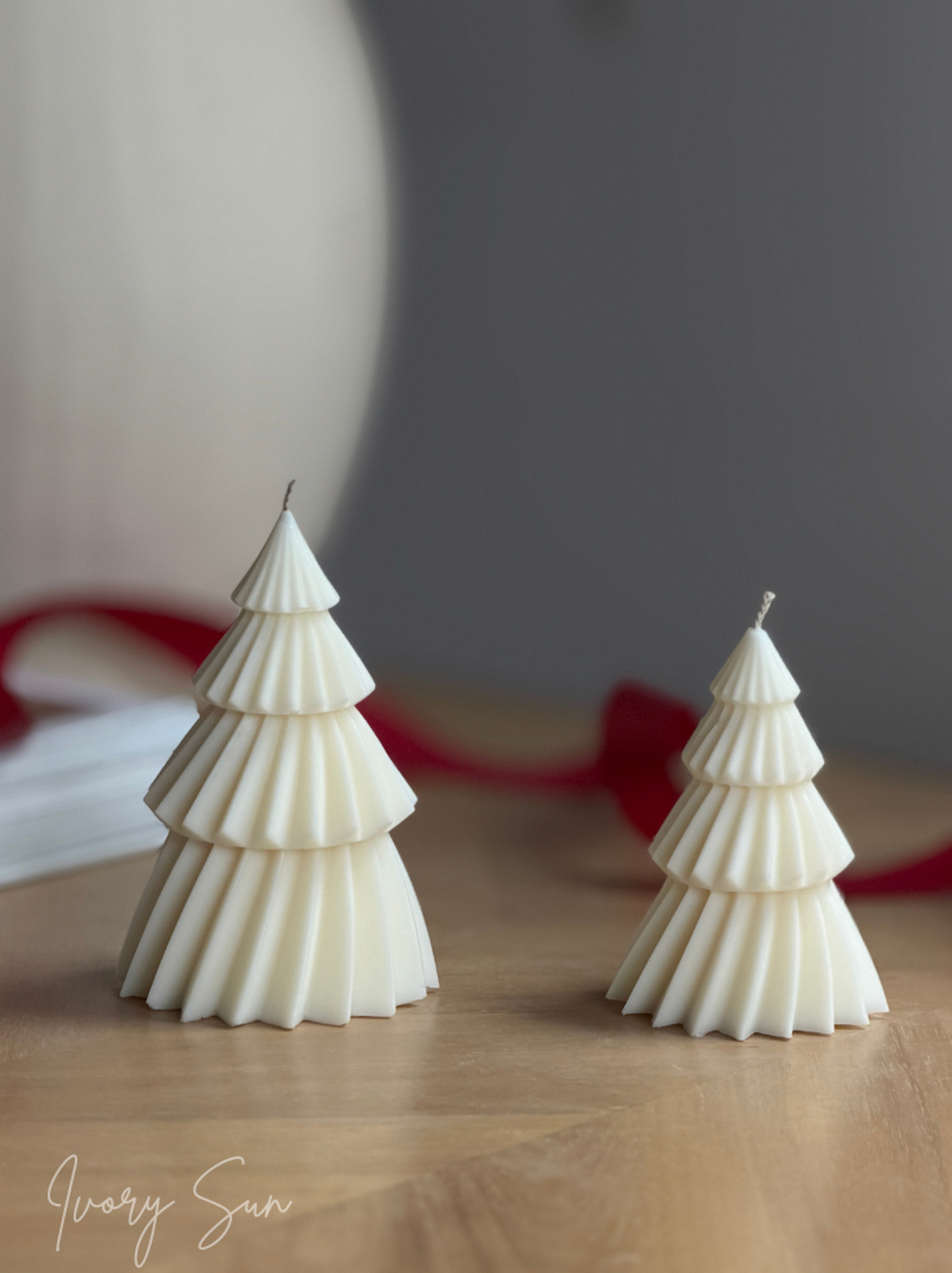 Two white soy Christmas tree shaped candles with pleated detailing, displayed on a light wooden surface with a soft-focus gray background and a red ribbon. Handmade Christmas candles by Ivory Sun, beautifully presented as a Christmas gift set. Discover Christmas gift ideas and Christmas Candles in Australia.