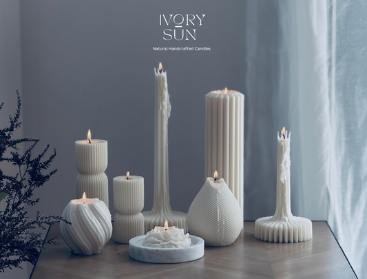Candle gift cards. A collection of white handcrafted soy candles in various sculptural shapes and heights, styled on a wooden table against a gray wall with a curtain. The brand name 'IVORY SUN' is displayed at the top. Ivory Sun gift cards available online.