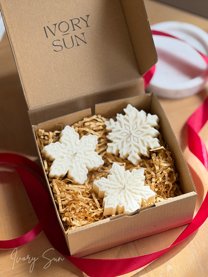 Three white soy snowflake candles displayed in a brown gift box filled with shredded paper, with the brand name 'IVORY SUN' printed inside the lid and a red ribbon around the box. Handmade Christmas candles by Ivory Sun, beautifully presented as a Christmas gift set. Discover Christmas gift ideas and Christmas Candles in Australia.