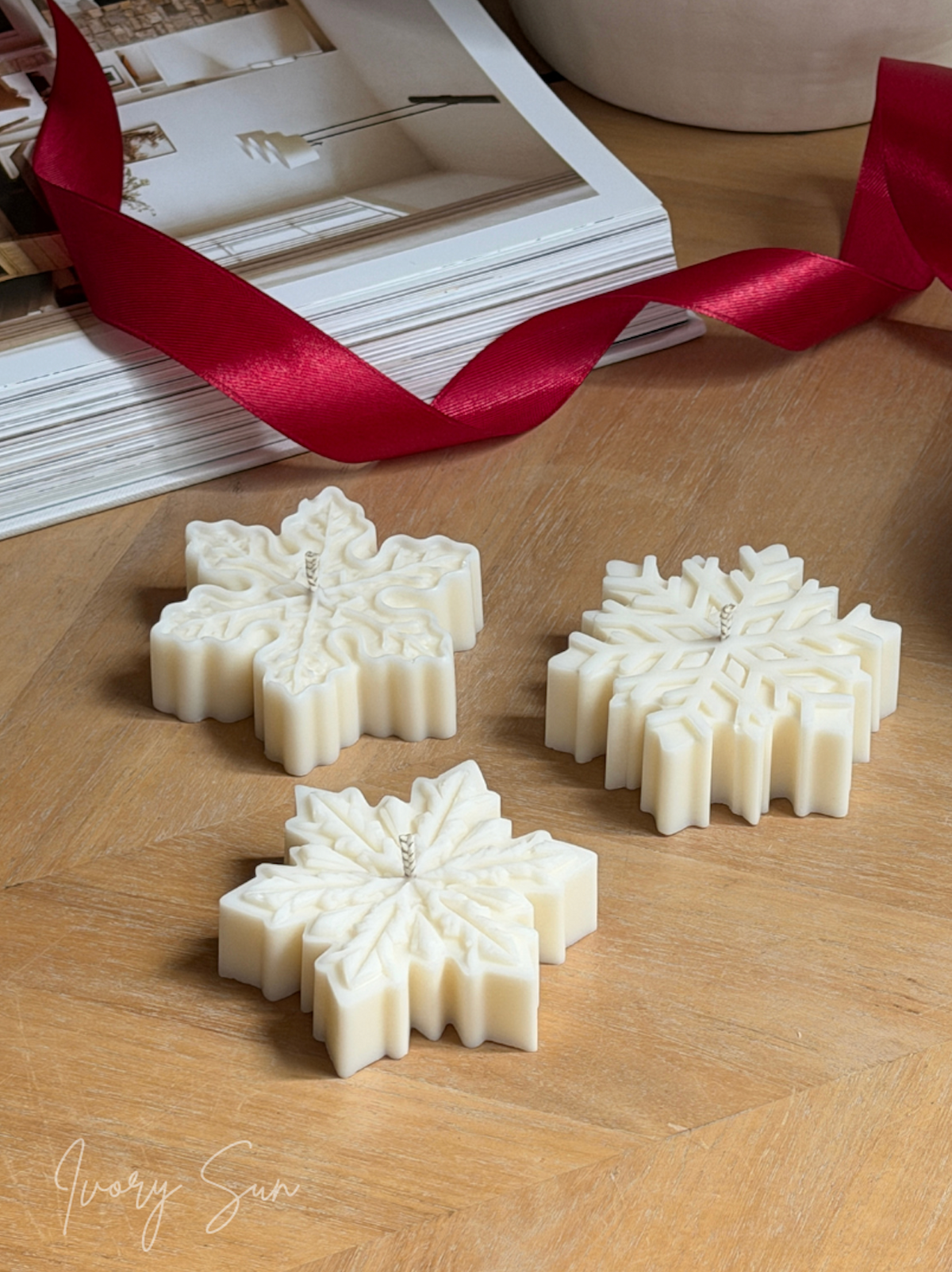 Three soy snowflake candles arranged on a wooden tabletop, with a red ribbon draped across the background and a book behind them. The brand name 'Ivory Sun' is visible in the bottom left corner. Handmade Christmas candles by Ivory Sun, beautifully presented as a Christmas gift set. Discover Christmas gift ideas and Christmas Candles in Australia.