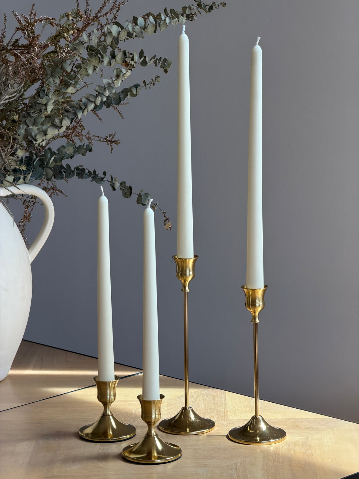 Four white taper candles in gold metal candlesticks arranged on a light wood surface against a muted gray wall, with dried greenery and part of a white ceramic vase visible. Handmade taper candles by Ivory Sun.