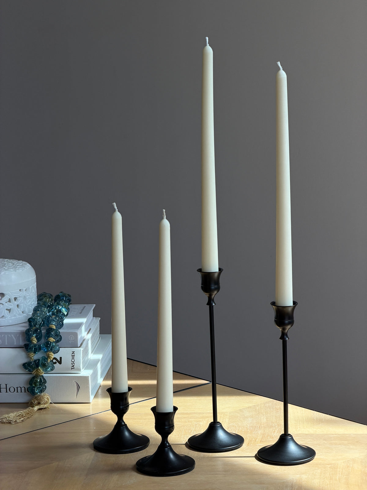 Four black metal taper candle holders of varying heights arranged on a light wood tabletop against a dark gray wall, with decorative books and a blue beaded garland on the left. Handmade taper candles by Ivory Sun.