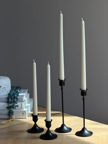 Four black metal taper candle holders of varying heights arranged on a light wood tabletop against a dark gray wall, with decorative books and a blue beaded garland on the left. Handmade taper candles by Ivory Sun.