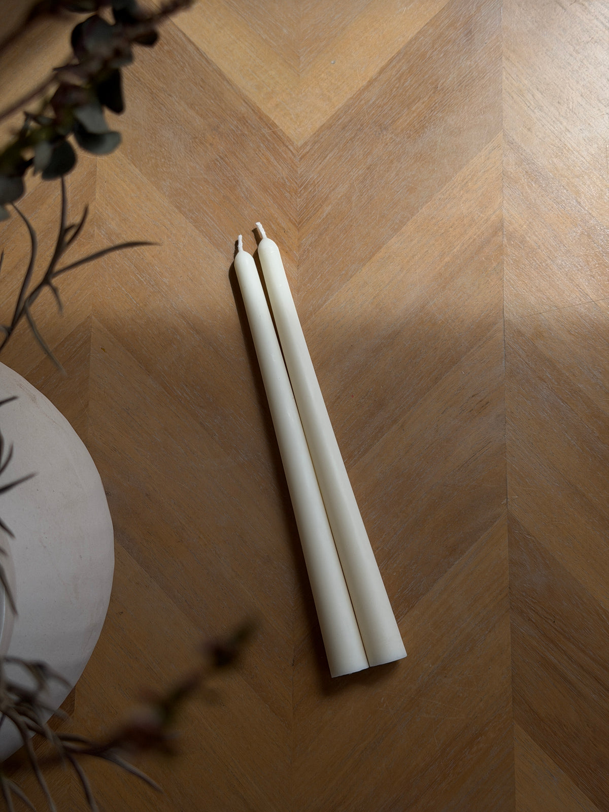 Two unlit white taper candles placed on a warm wood chevron floor, with soft shadows and decorative dried branches in the foreground. Handmade taper candles by Ivory Sun.