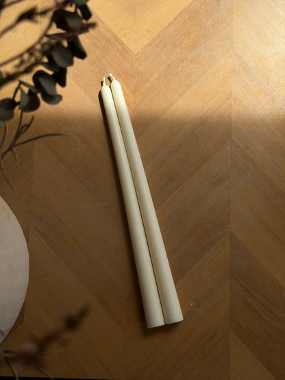 Two cream-colored taper candles placed on a warm wood herringbone floor, with soft shadows and dried plant stems in the background. Handmade taper candles by Ivory Sun.
